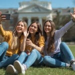 Three happy young women taking a selfie with a refurbished smartphone while sitting on green grass.